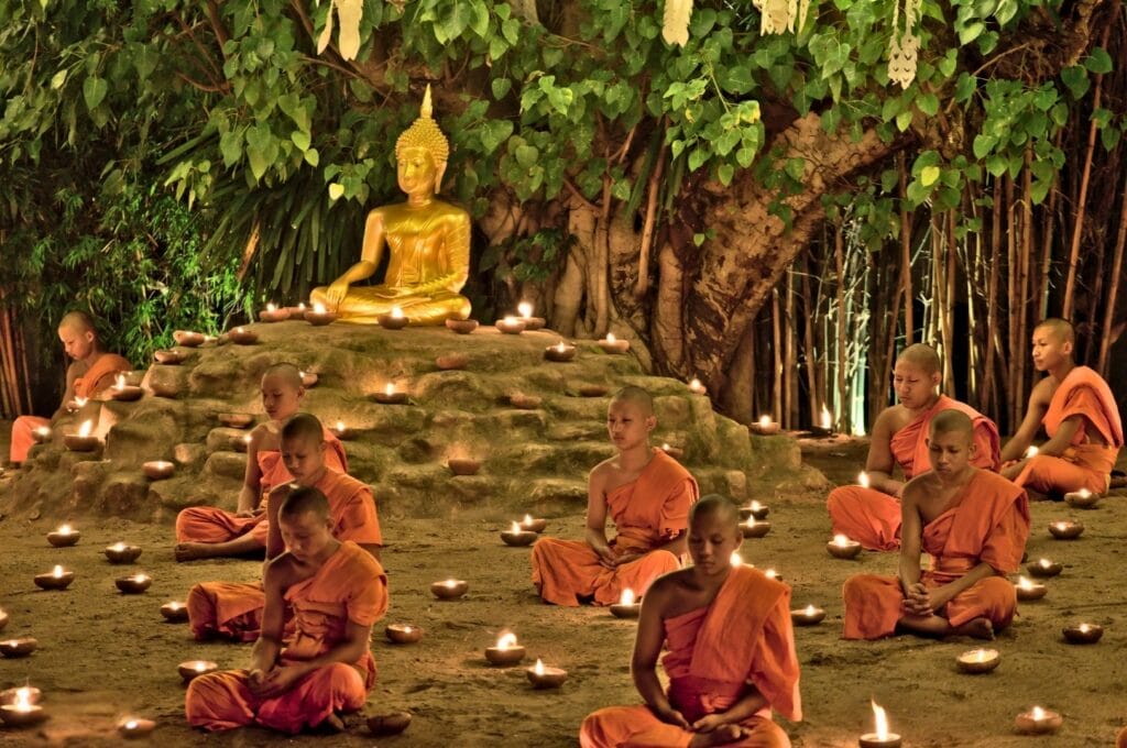 monks seated cross-legged around a golden Buddha statue in front of bamboo trees for a ceremony during the Festival of Lights.